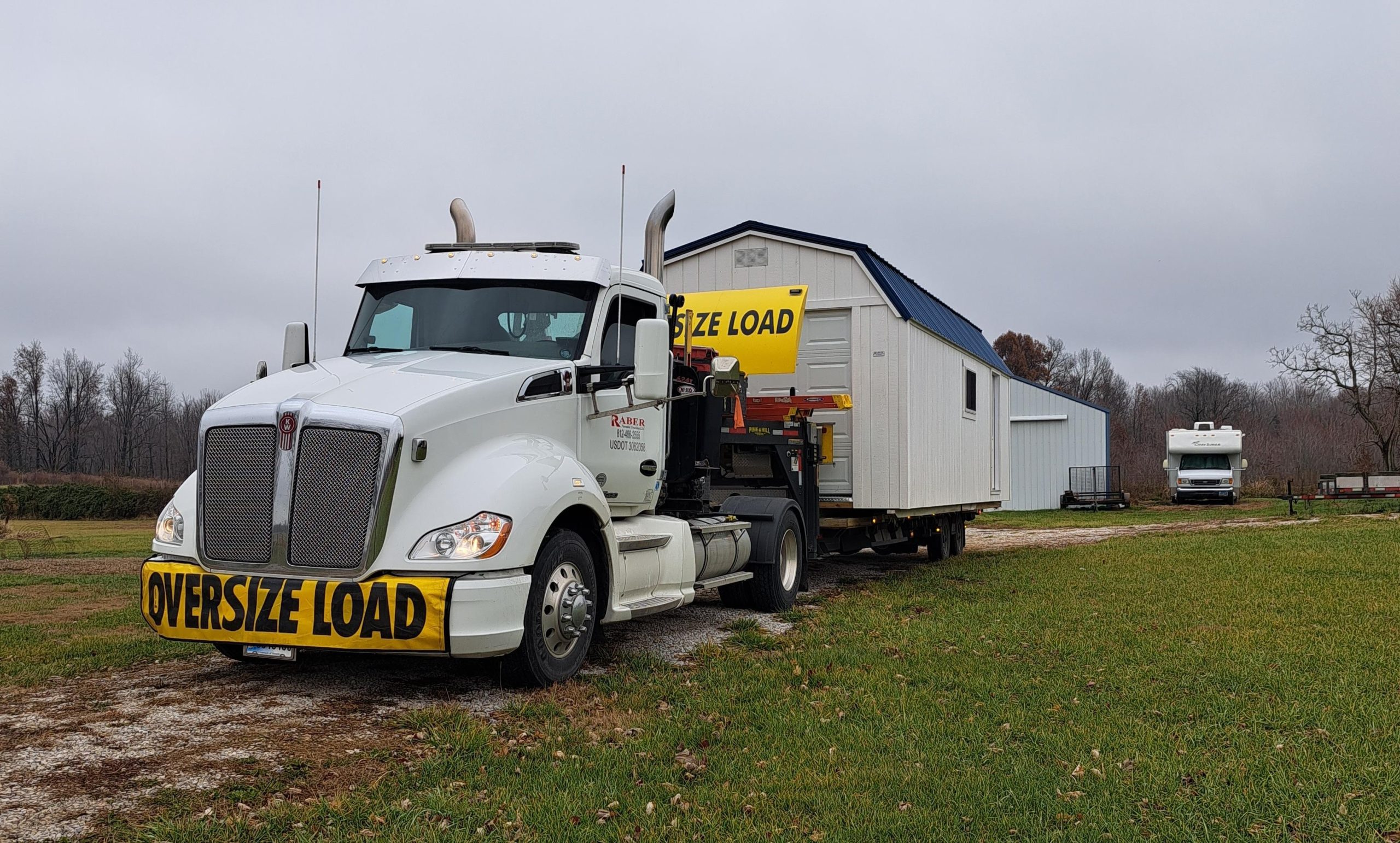 White truck with oversize load signs delivering a white storage shed to a property.