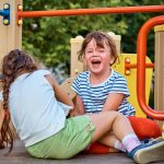 2 children sitting on an outdoor playset and laughing together.