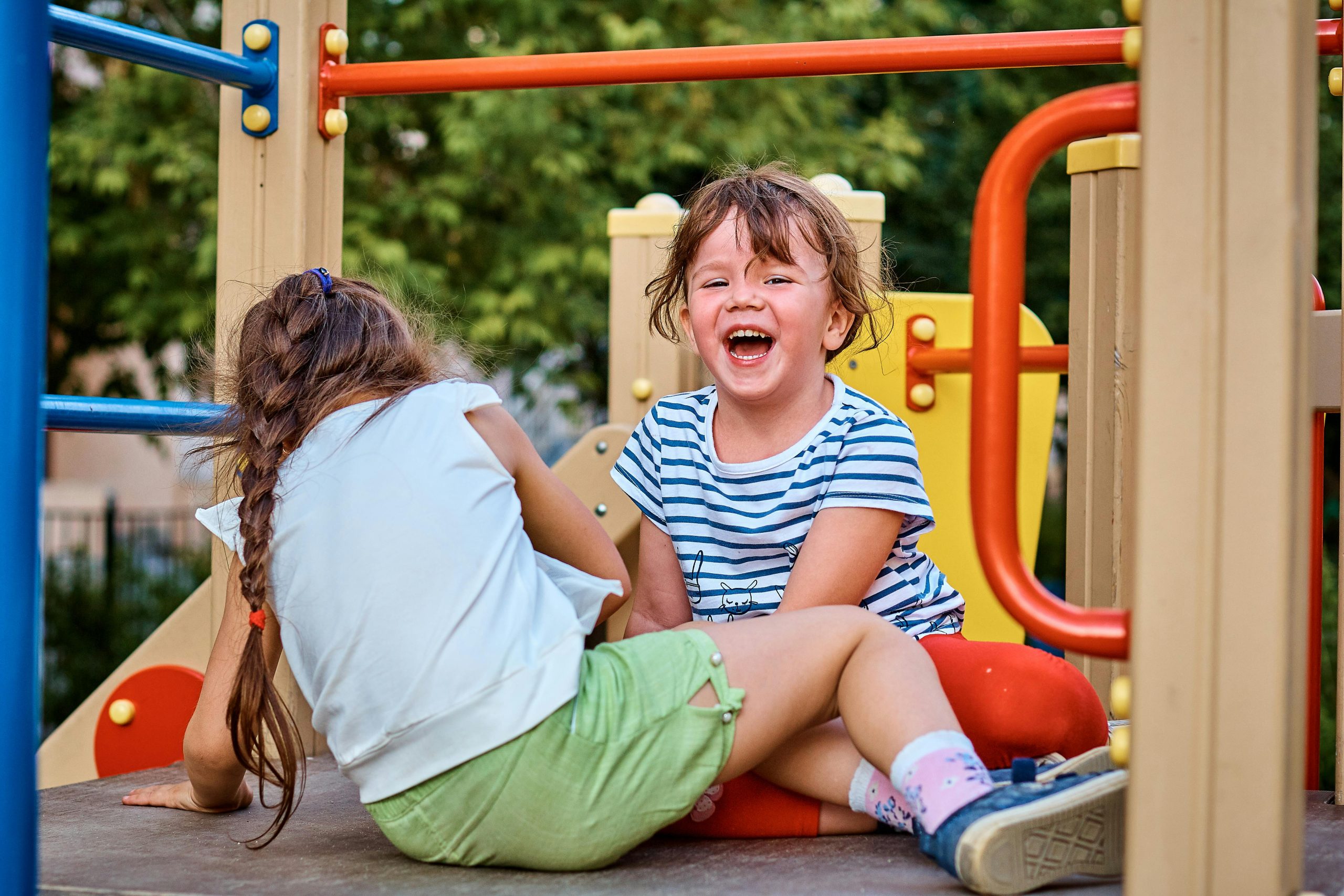 2 children sitting on an outdoor playset and laughing together.