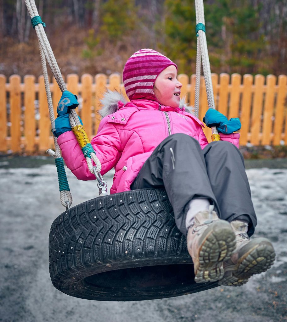 Child in a pink winter coat, pink winter hat, and dark gray snow pants swings on a tire swing.